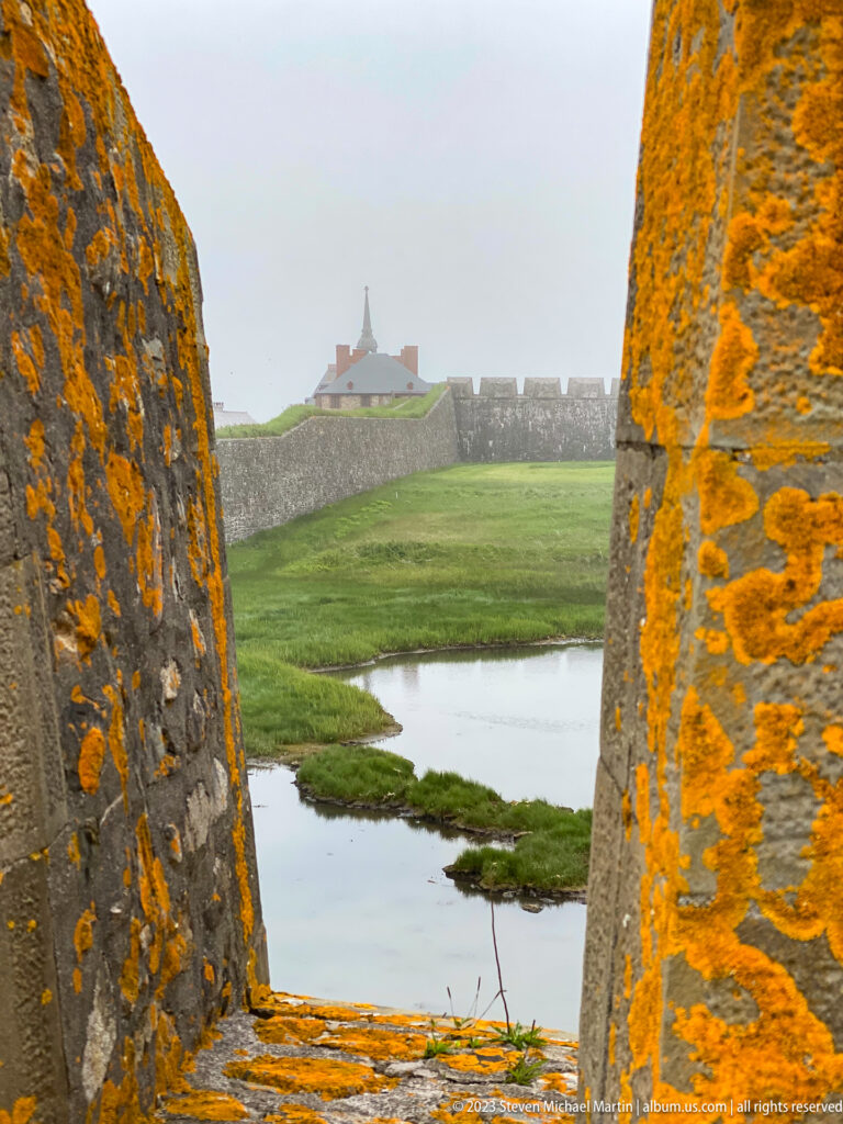 Water and grassy earthworks seen through a narrow opening in thick lichen-covered stone fortification.
