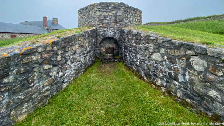 Narrow grassy stone passage leading to a small arched opening beneath a rounded masonry structure.
