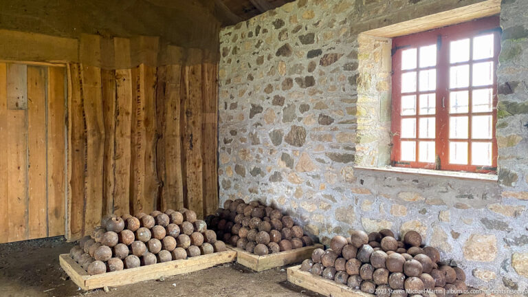 Pyramidal stacks of dark iron cannonballs rest on wooden platforms beside a stone wall and window.