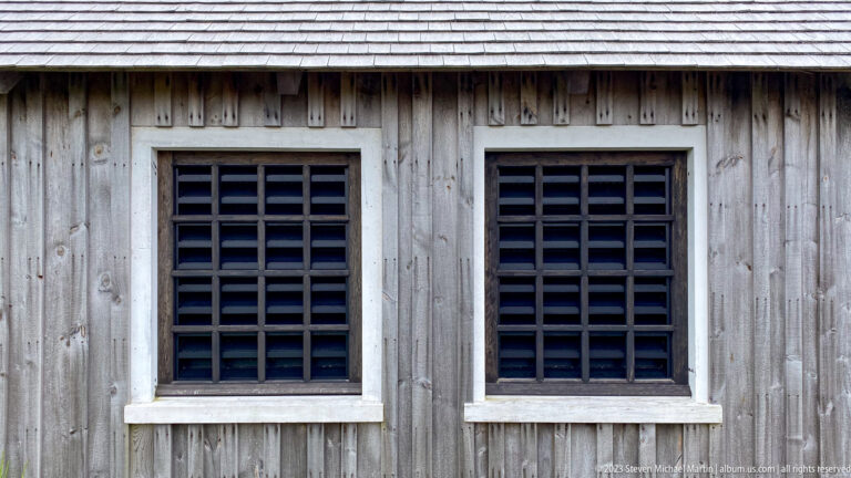 Two dark gridded windows set in weathered vertical wooden siding beneath a shingled roof.