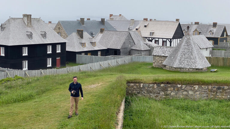 Visitor walks on grass below clustered timber and stone buildings, fences, and a conical-roofed outbuilding.
