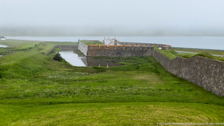 Stone fortress walls, water-filled ditch, and broad grassy earthworks extend toward a projecting defensive work.