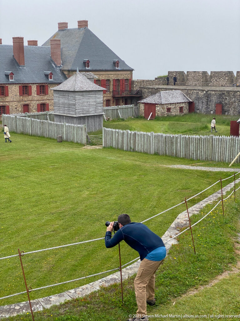 Visitor bends with a camera beside the parade ground, with barracks, fences, and stone defenses behind.