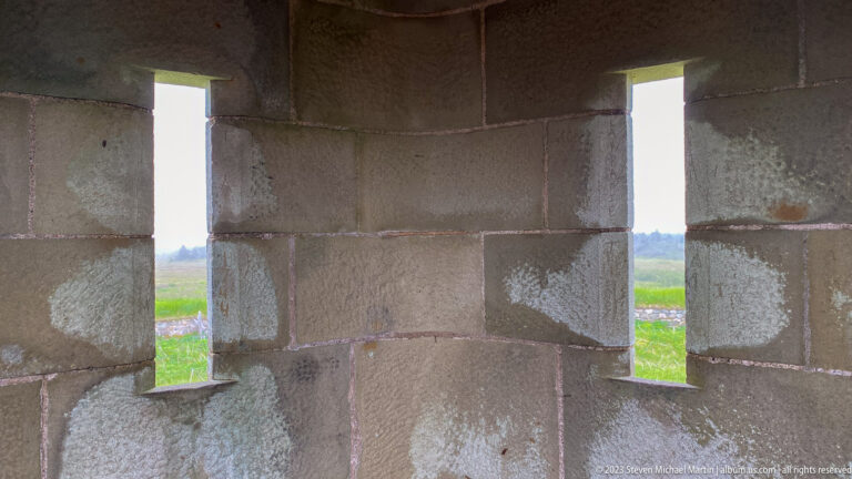 Two narrow vertical firing openings pierce a thick dressed-stone wall, showing grass outside.