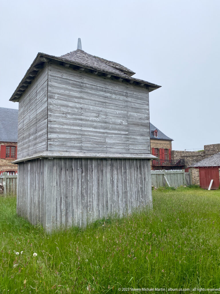 Small weathered wooden outbuilding with vertical boards and a low hipped shingle roof.