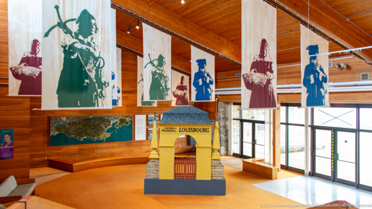 Visitor centre gallery with hanging portrait graphics, a map, and a model of a Louisbourg gate.