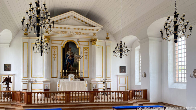 White-and-gold chapel sanctuary with central devotional image, altar, chandeliers, and wooden rail.