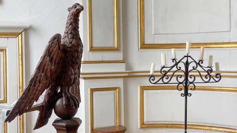 Dark carved wooden eagle with spread wings beside a wrought-iron candle stand in a chapel.