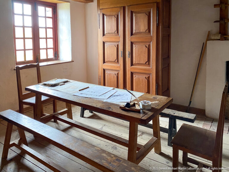 Long wooden table with papers and writing tools, benches, window, and tall paneled cupboard in a historic room.