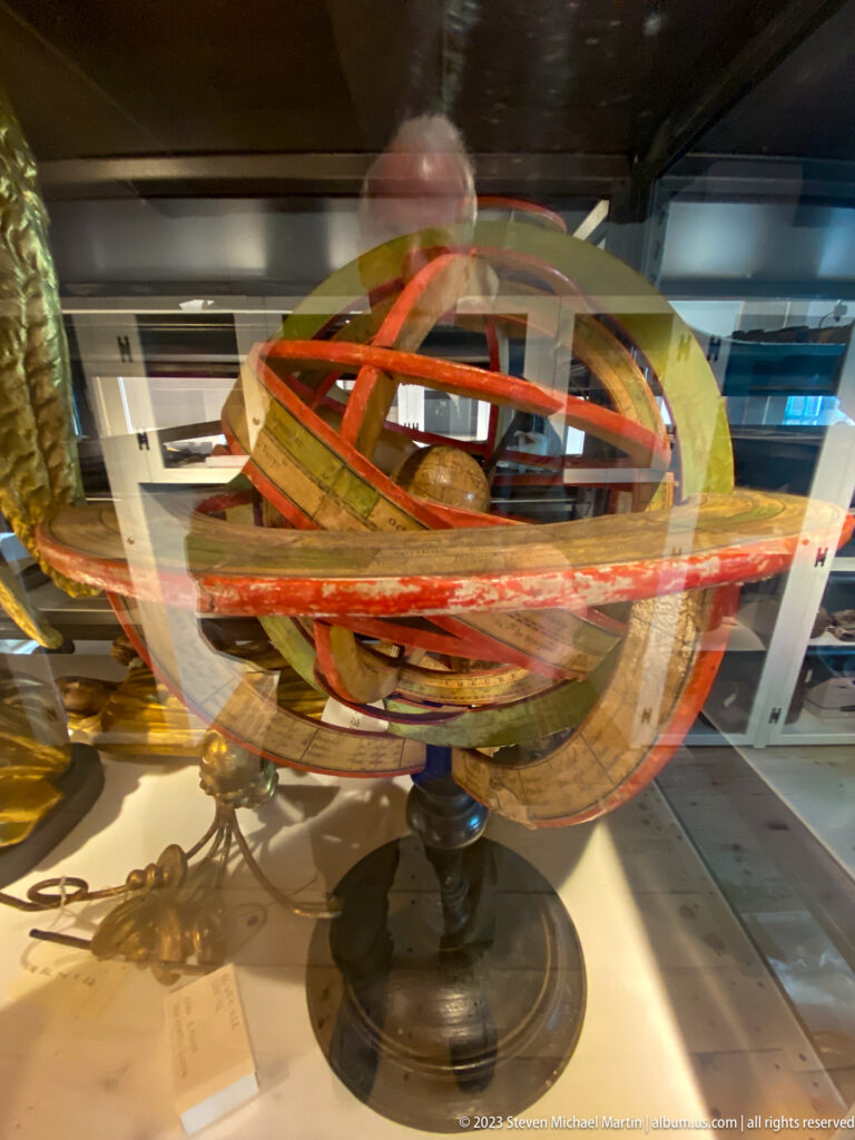 Painted armillary sphere with intersecting rings mounted on a dark pedestal inside a glass display case.