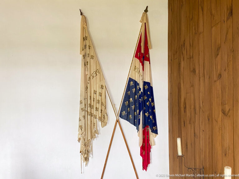 Two historic-style flags on crossed poles, including blue, red, white, and fleur-de-lis designs, displayed indoors.