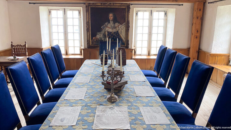 Long patterned table with blue chairs, candlesticks, papers, tall windows, and a portrait in a formal historic room.