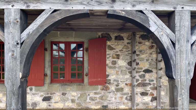Red-shuttered multi-pane window in rough stone masonry beneath weathered curved timber braces.