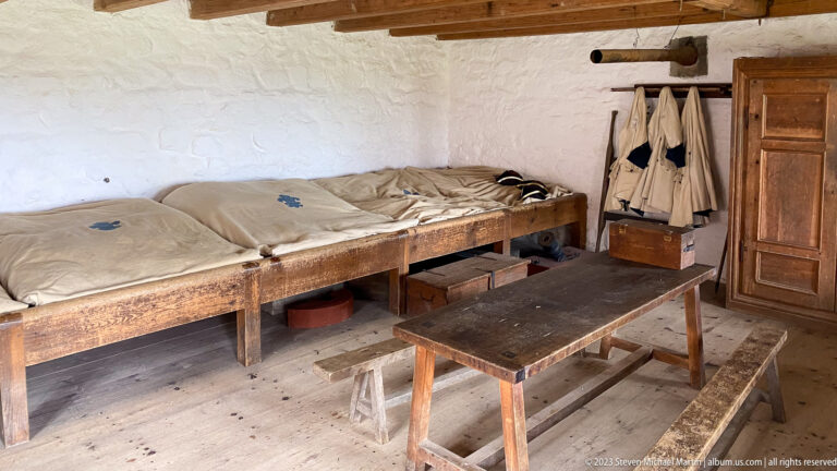 Wooden sleeping platforms with blankets, storage boxes, hanging garments, benches, and table in a whitewashed barracks room.