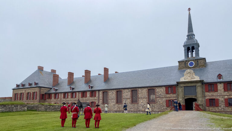 Four red-coated interpreters walk across grass before the long stone barracks and central chapel spire.
