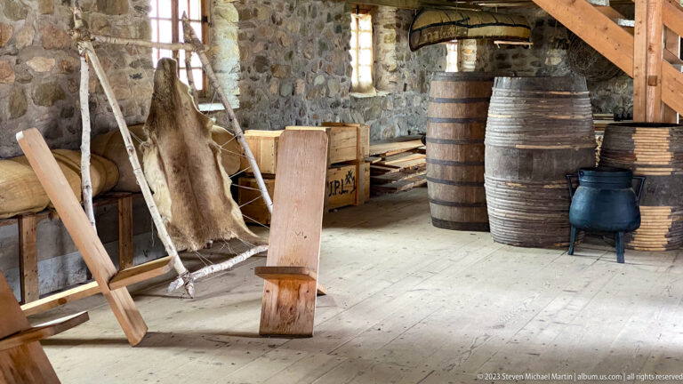 Stone storehouse with wooden barrels, animal hides, crates, planks, and a black iron vessel.