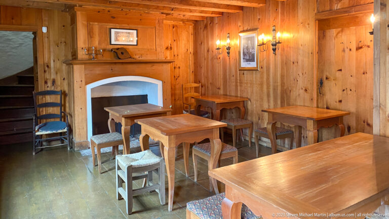 Warm wooden interior with small tables, stools, chairs, candle sconces, and a white-plastered hearth opening.