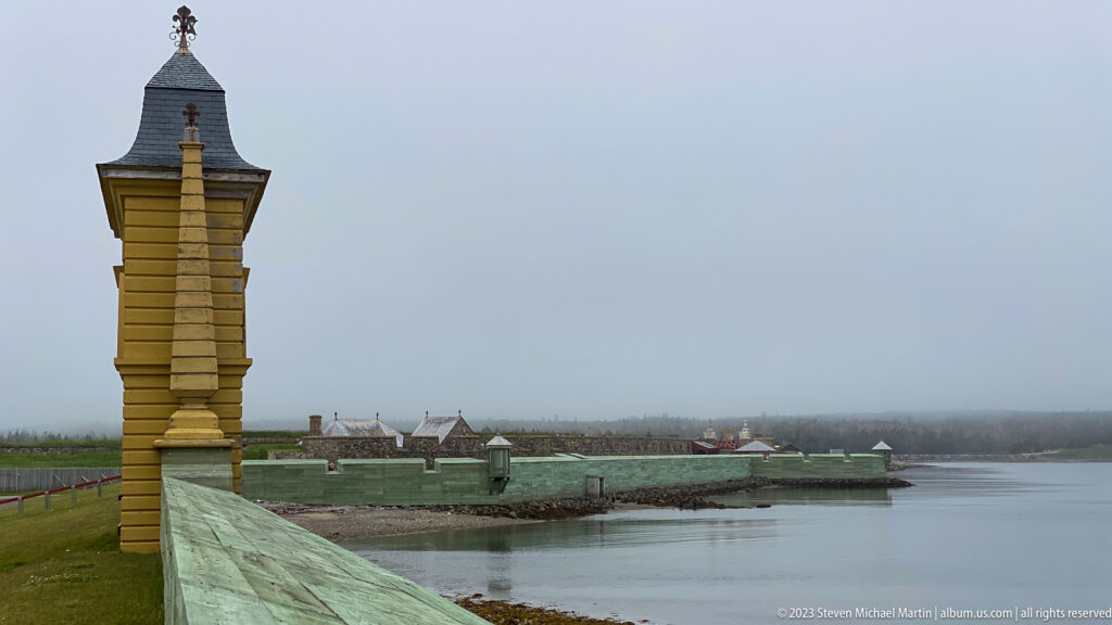 Tall wooden sentry structure beside a green parapet overlooking calm water and distant Louisbourg fortifications.