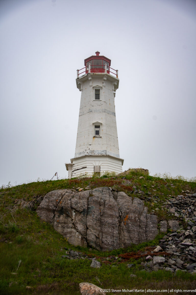 White tapered lighthouse with red lantern room standing on exposed coastal rock and low vegetation.