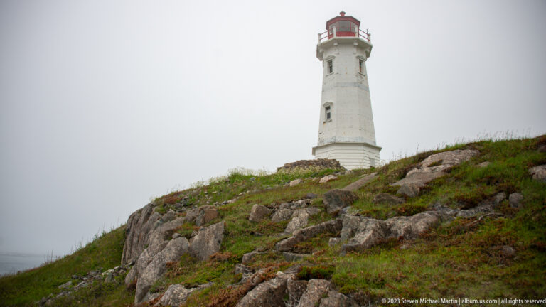 White Louisbourg Lighthouse with red lantern room viewed from below across rock and coastal vegetation.