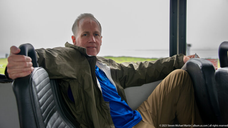 Male visitor in blue shirt and dark jacket seated sideways inside a shuttle bus.