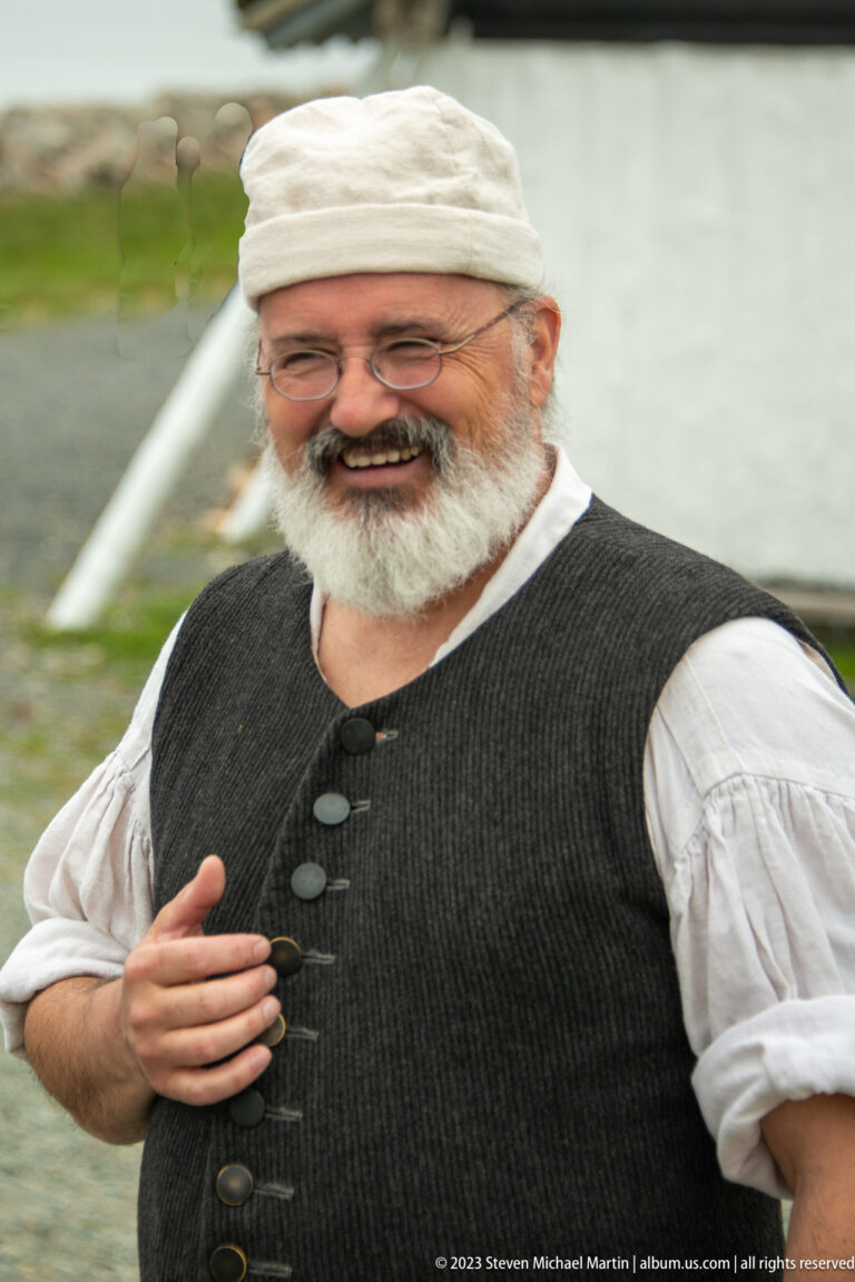 Bearded historical interpreter smiling in a linen cap, white shirt, and dark waistcoat.