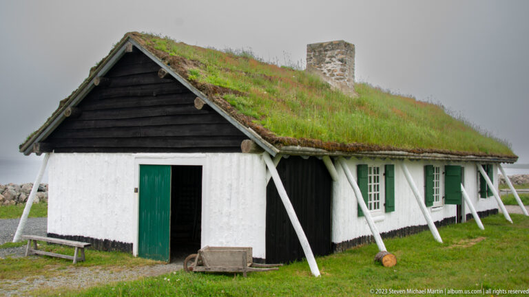 Whitewashed reconstructed house with dark gable, green shutters, chimney, and grass-covered roof.