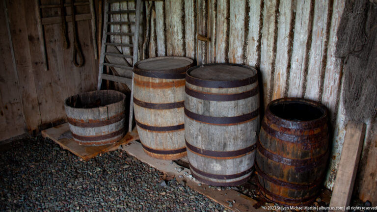 Group of weathered wooden barrels and tubs with iron hoops inside a rough-board storage room.