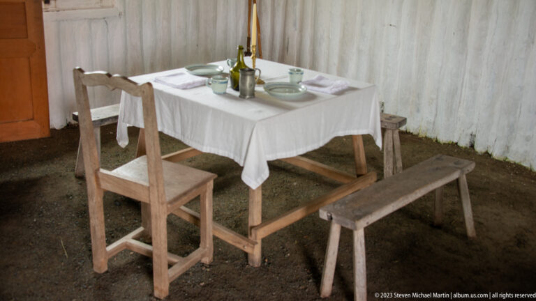 Small cloth-covered table set with ceramics, glasses, candles, chair, and wooden bench.