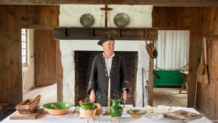 Costumed male interpreter standing behind a table in a reconstructed room with a large white hearth.