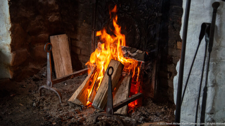 Bright wood fire burning in a soot-blackened masonry hearth with iron tools beside it.