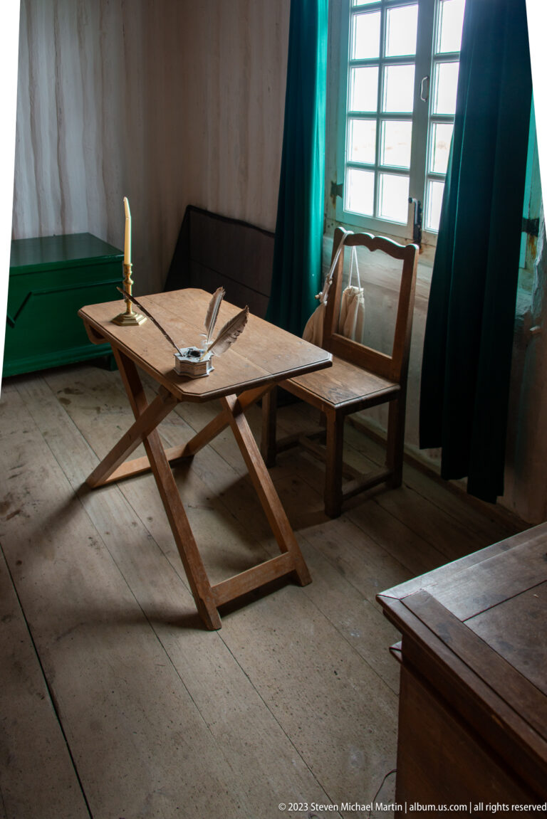 Wooden writing table with papers, quill, and candle beside a bright window in a sparse room.