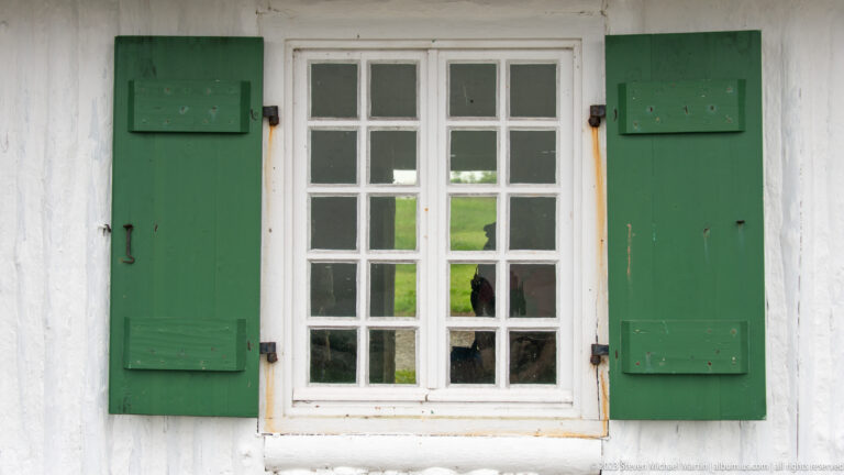 White multi-pane window flanked by weathered green wooden shutters on a white wall.