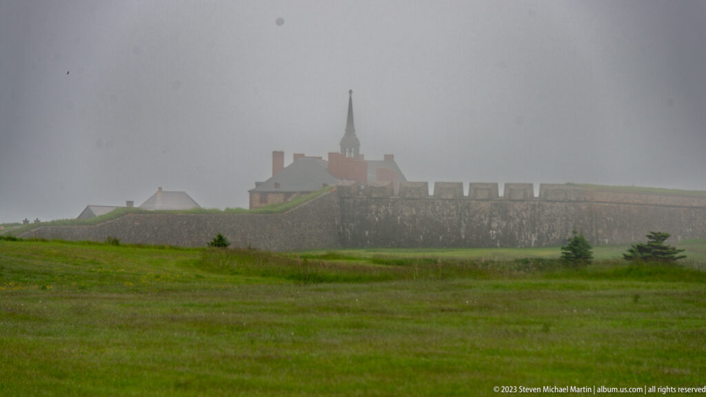 Foggy distant view of stone fortifications, roofs, and a church tower across green grass.