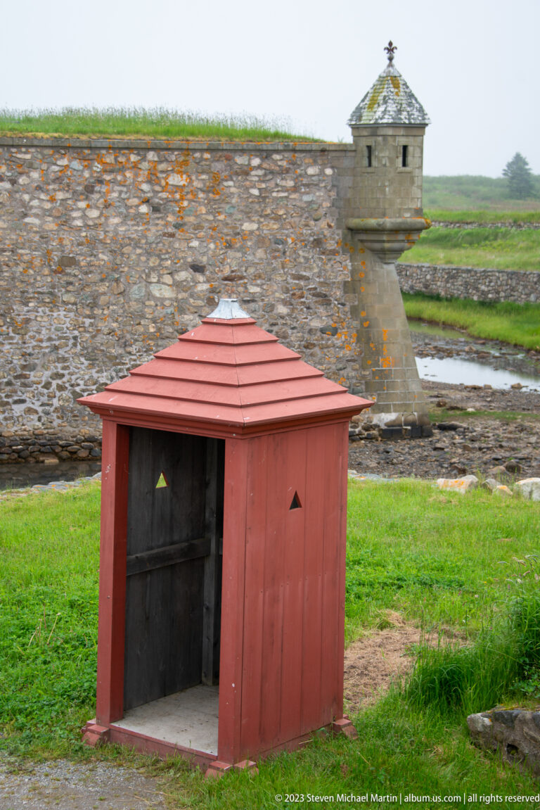 Small red wooden shelter in front of gray stone walls and a projecting sentry structure.