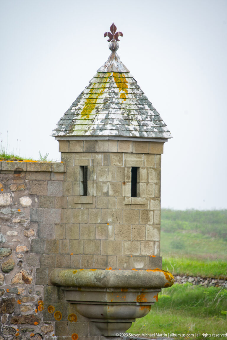 Small stone sentry box with lichen-covered shingle roof, narrow openings, and a metal weather vane on the fortress wall.