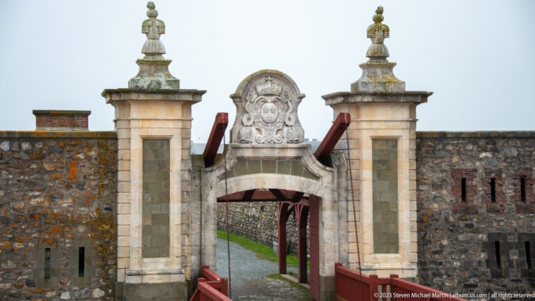 Close frontal view of a stone fortress gateway with crowned fleur-de-lis shield, scrollwork, and flanking masonry piers.
