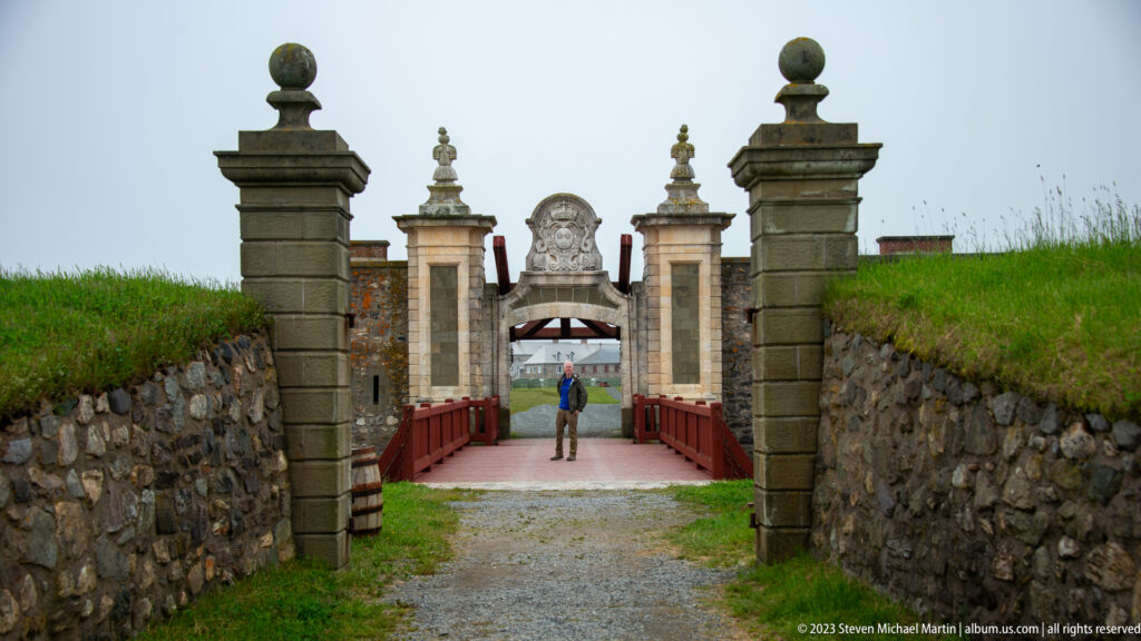 Visitor walking toward an ornate fortress gate and red bridge, seen between dark stone posts and grassy embankments.