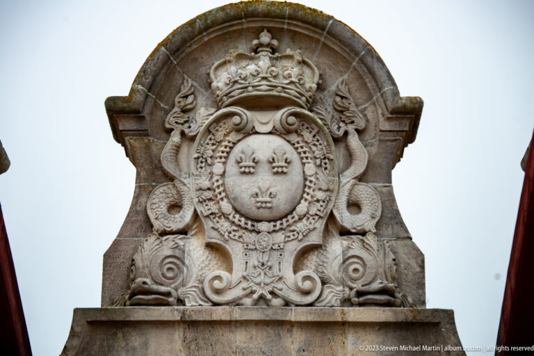 Carved stone royal arms with crown, three fleurs-de-lis, chains, scrollwork, and ornamental relief above the fortress gate.