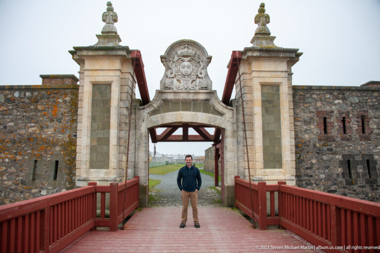 Visitor standing on a red wooden bridge beneath an ornate stone gateway with crowned heraldic carving and fortress walls.