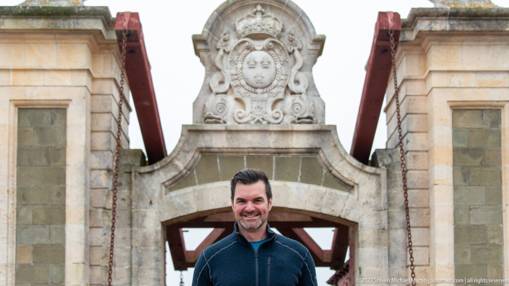 Smiling male visitor framed by pale stone gate piers beneath a crowned fleur-de-lis heraldic carving.