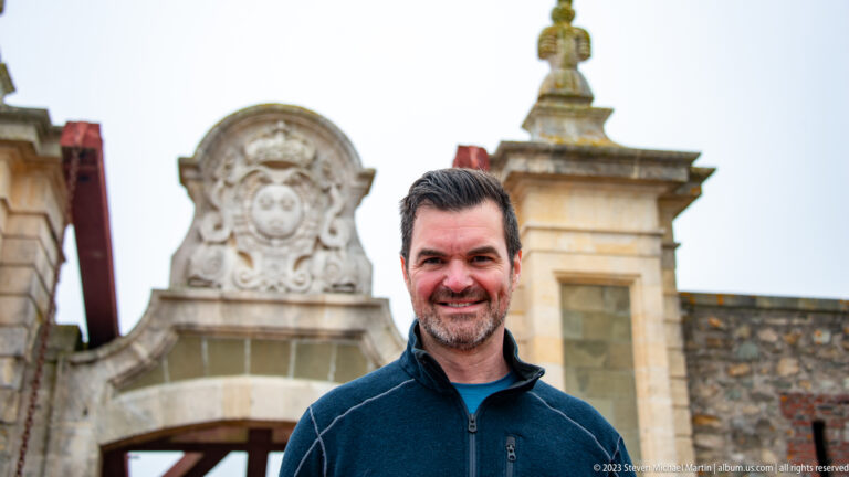 Close portrait of a smiling male visitor with the crowned fortress gateway and pale stone pier blurred behind him.