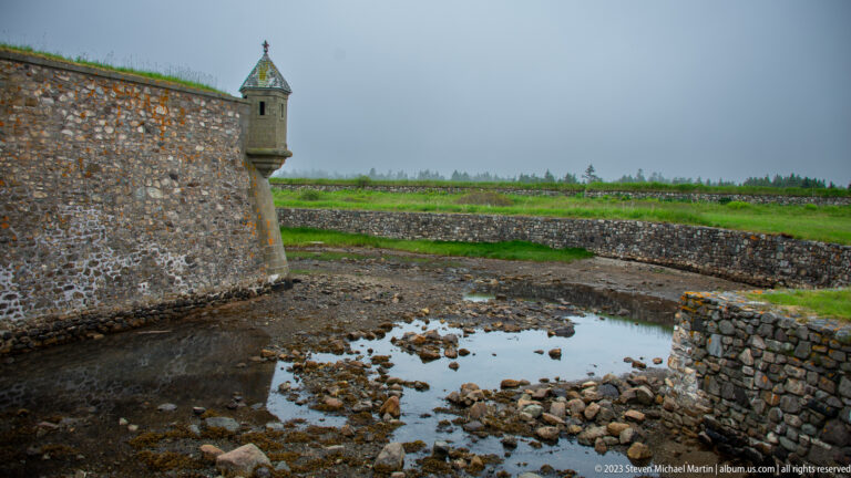 Angular stone fortress wall beside a shallow rocky ditch, with grassy earthworks and a small roofed lookout in fog.