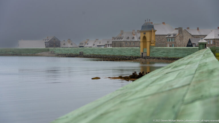 Mist-covered reconstructed fortress town across calm water, with a yellow waterfront structure and pale green foreground roof or rampart.