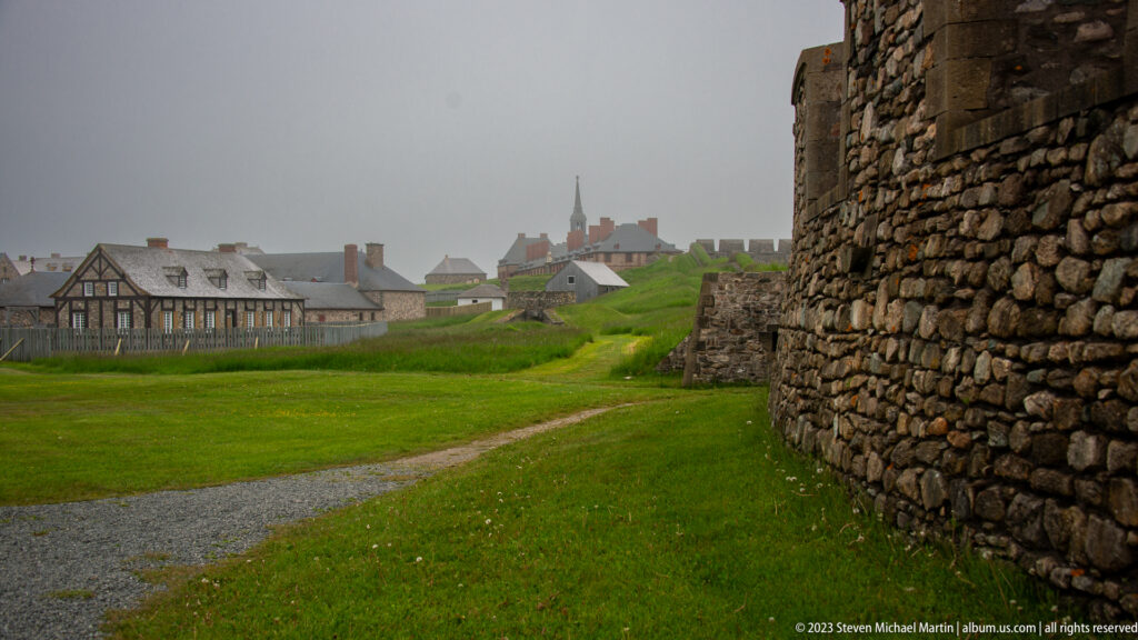 Grassy lane beside a rough stone fortress wall, with timber-framed buildings and a distant church spire in mist.