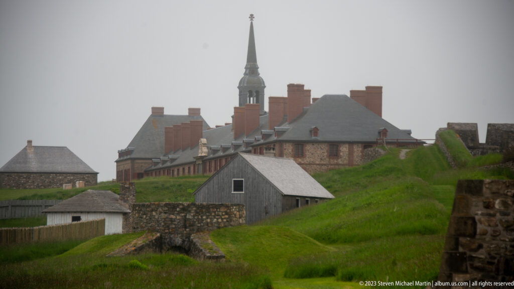 Clustered reconstructed stone and timber buildings with many chimneys and a tall church spire behind grassy earthworks.