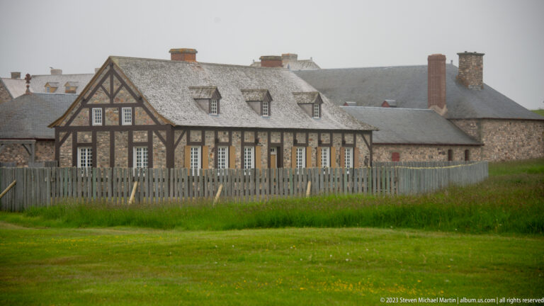 Long half-timbered building with dormer windows, steep shingle roof, chimneys, and a pale wooden fence in front.