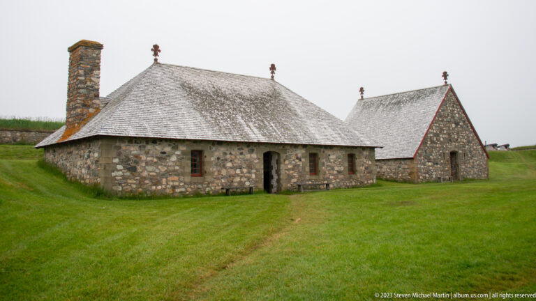 Two low stone buildings with steep shingle roofs on grass, the nearer one with a tall chimney and red-trimmed openings.