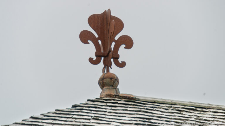 Large metal fleur-de-lis finial mounted on a wooden base above the ridge of a weathered shingle roof.