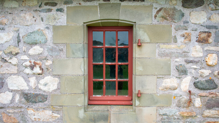 Red multi-pane window framed in dressed stone within a rough fieldstone wall, with small iron fittings beside the opening.